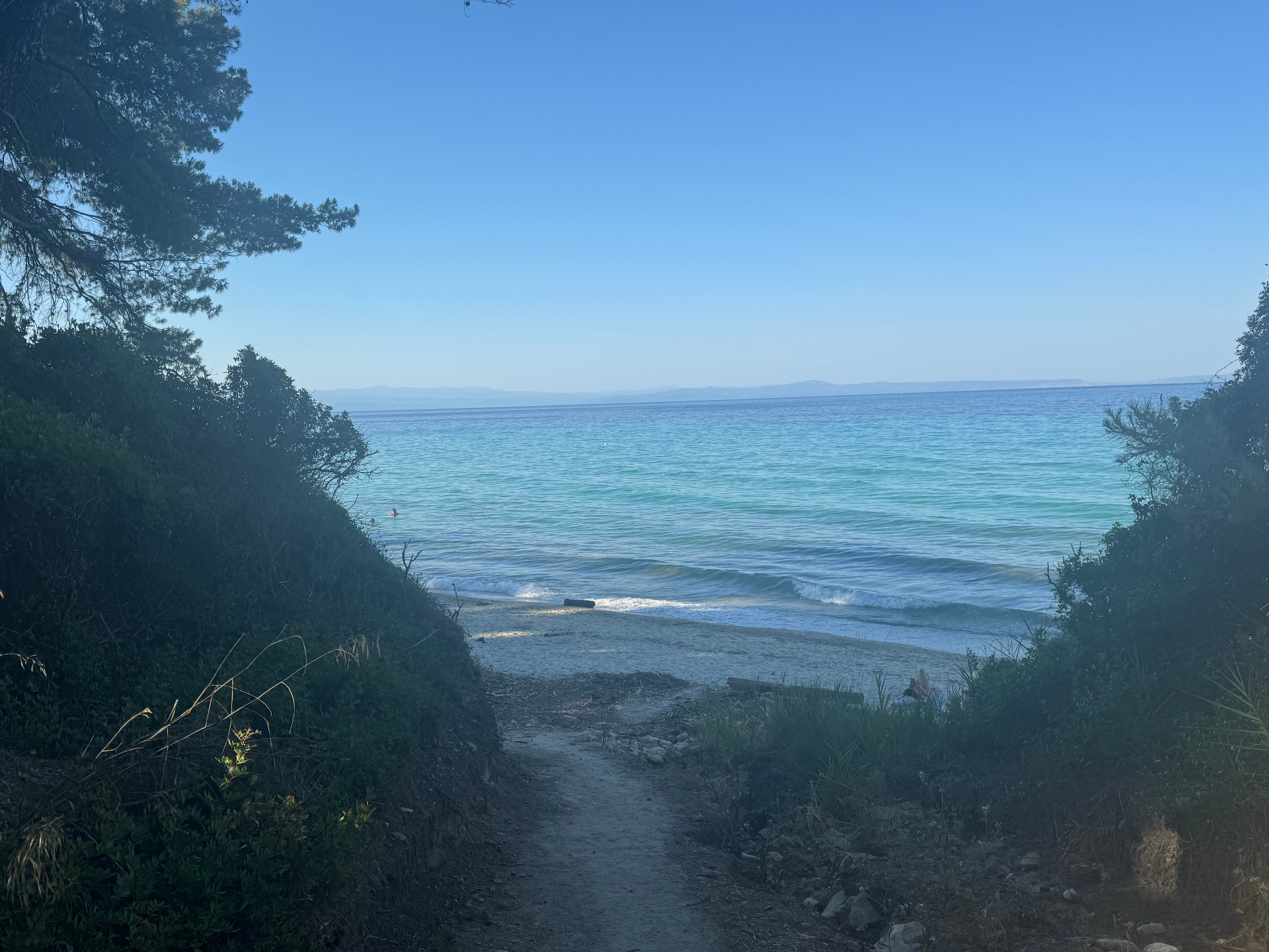 A sandy footpath descending through dense maquis shrubland to the turquoise shallows of a Kriopigi cove.